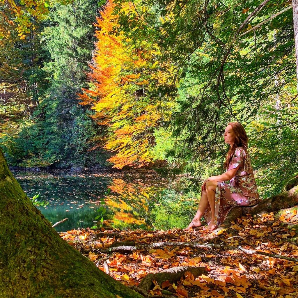 Denise Mayer sitzt an einem See im Herbstwald auf einem Stein.