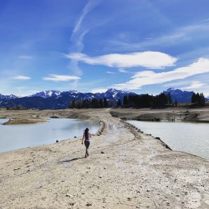 Denise läuft auf einer Sandbank weg von der Kamera. Links und Rechts von ihr ist Wasser. In der mitte geht ein Kies Weg hindurch. Der Himmel ist blau mit vereinzelten Wolken