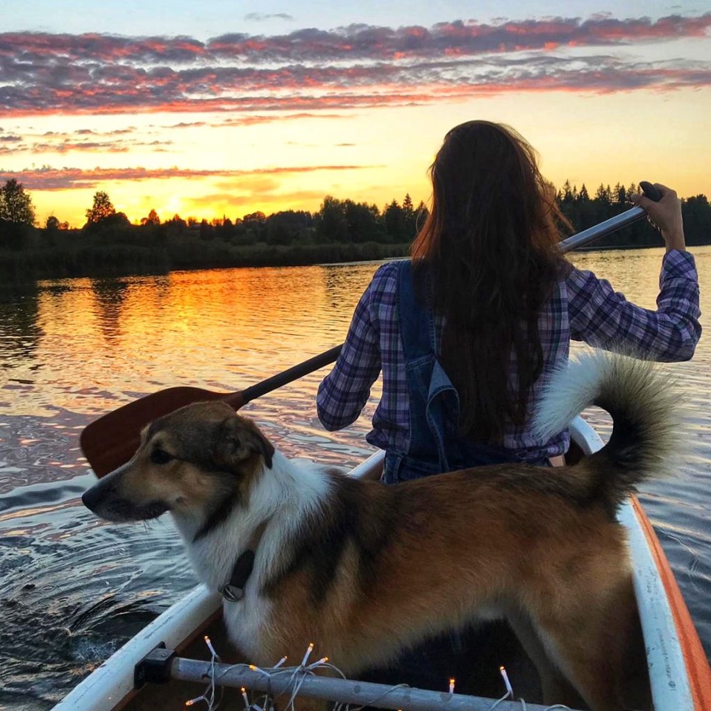 Denise sitzt in einem Boot mit einer Paddel in der Hand. Direkt hinter ihr steht ihr Hund der nach links sieht. In weiter Ferne sieht man einen leichten Sonnenuntergang mit gelb rötlichem Himmel.